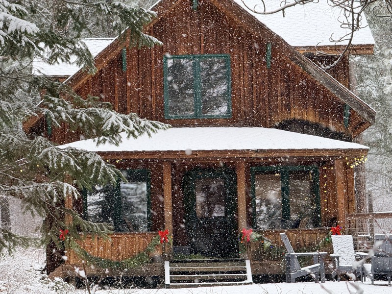 The cabin's covered porch dusted with fresh snow and decorated with holiday garland during a snowfall
