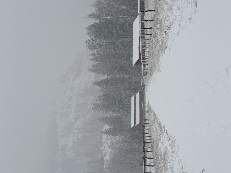 A snow-covered path leading between farm barns toward misty, forested mountains