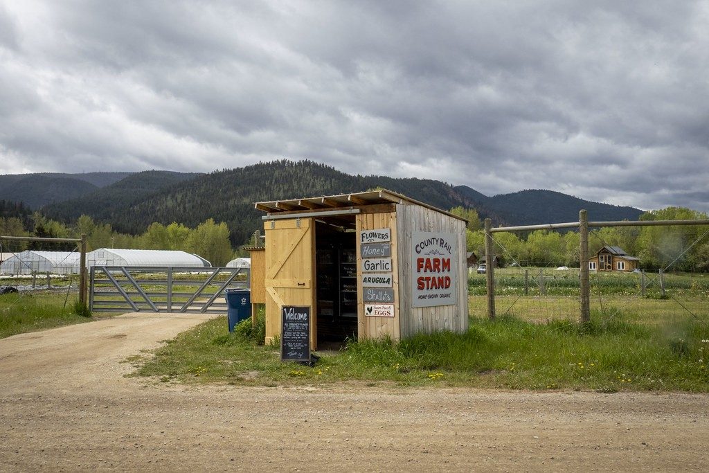 A roadside farm stand with hand-painted signs, greenhouses and mountains behind