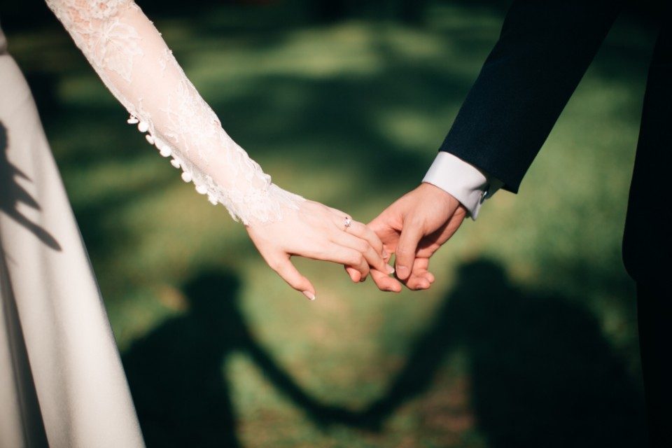 A newly married couple holding hands with a wedding ring, shadows cast on the grass